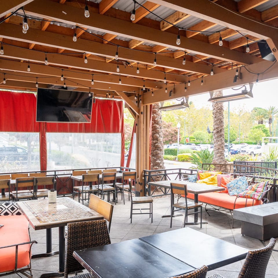Covered restaurant patio with string lights, wooden tables, colorful cushioned seating, red curtains, and palm trees outside.