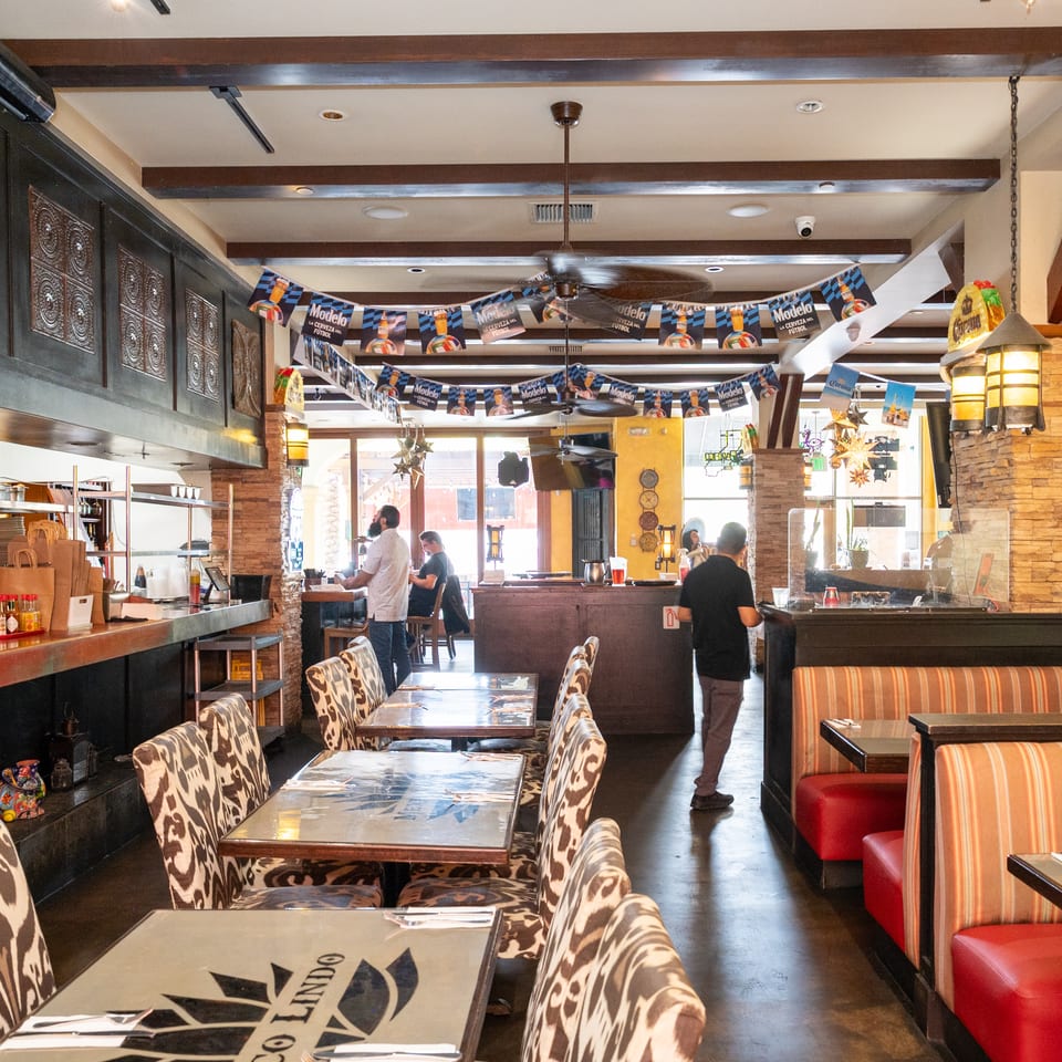 Spacious Mexican restaurant interior with patterned booths, dining tables, stone columns, ceiling fans, and Modelo beer decorations.