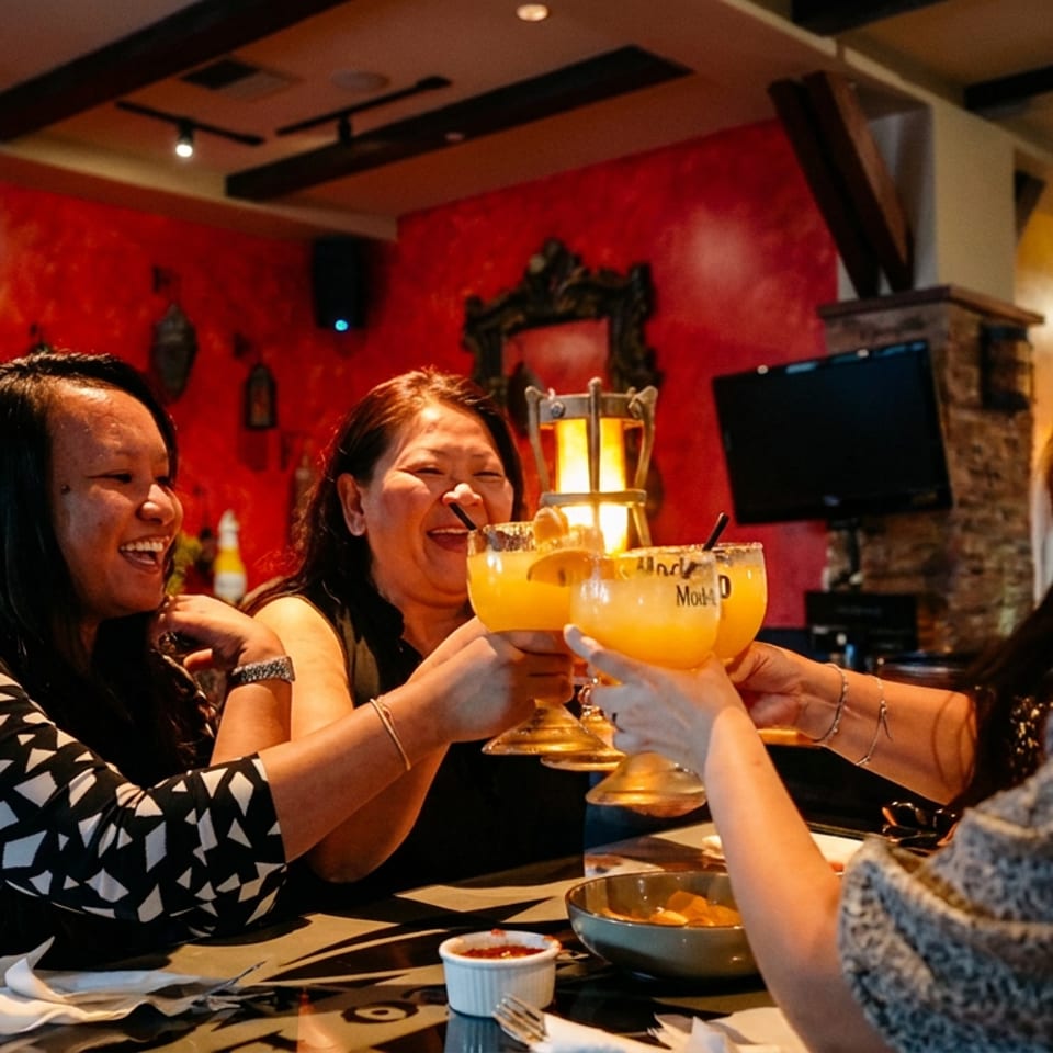 Three women laughing and toasting large margaritas in a vibrant, red-walled restaurant.