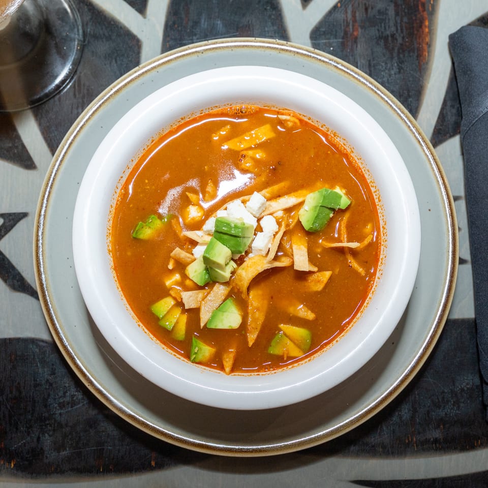Overhead view of spicy red soup topped with avocado, cheese, and crispy tortilla strips on a restaurant table.