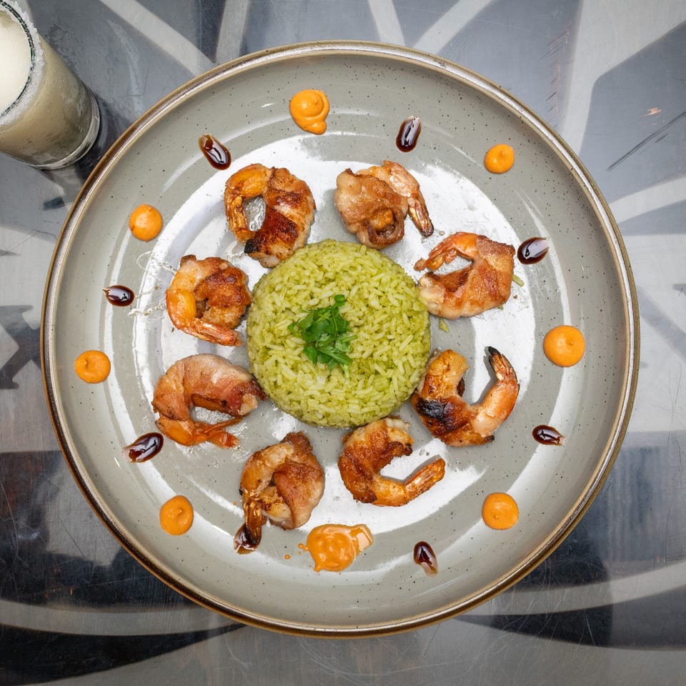 Overhead view of grilled shrimp surrounding green rice, drizzled with orange and dark sauces, alongside a salt-rimmed lime cocktail.