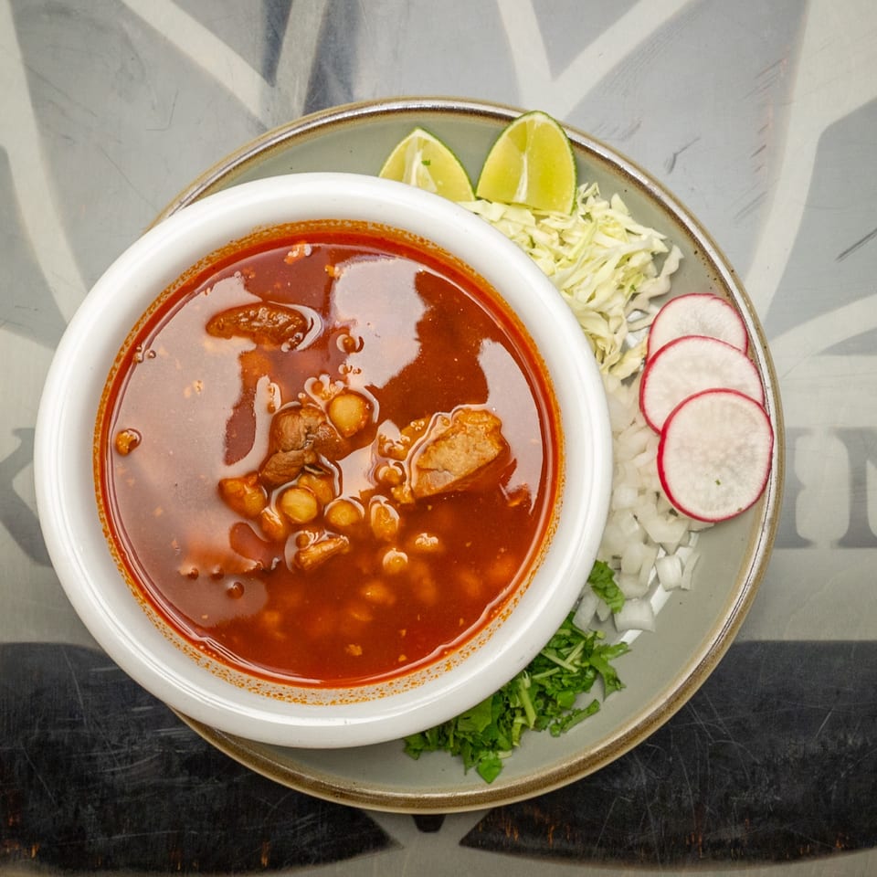 Overhead view of red pozole with hominy and meat, served with cabbage, radishes, lime wedges, onion, and cilantro.