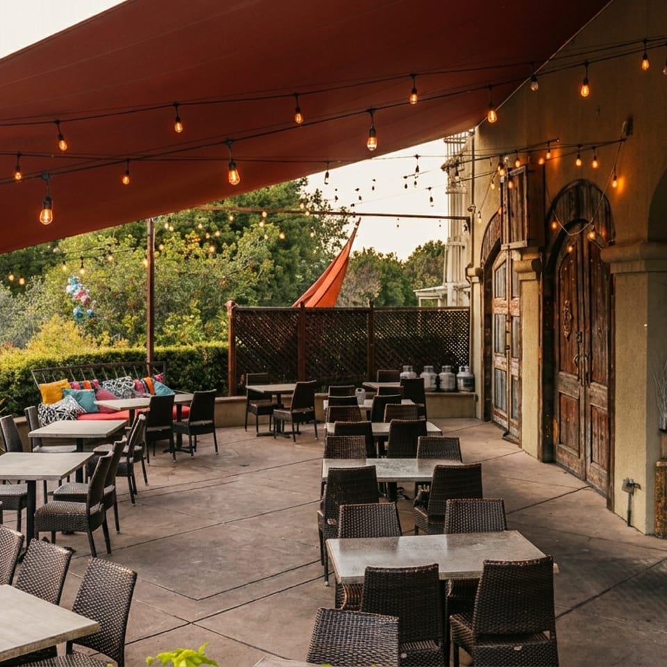 Outdoor restaurant patio with tables, chairs, string lights, and a red canopy, surrounded by lush greenery.