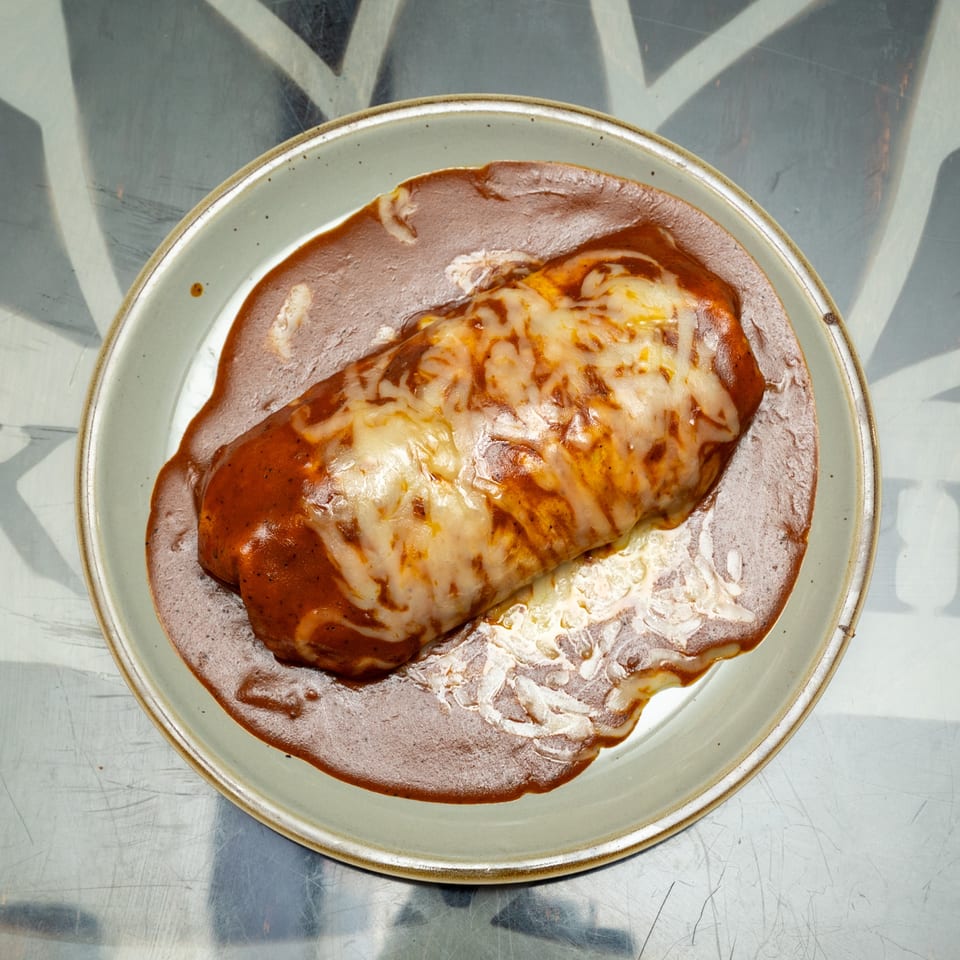 Cheesy enchilada smothered in red sauce on a ceramic plate atop a reflective metal table.