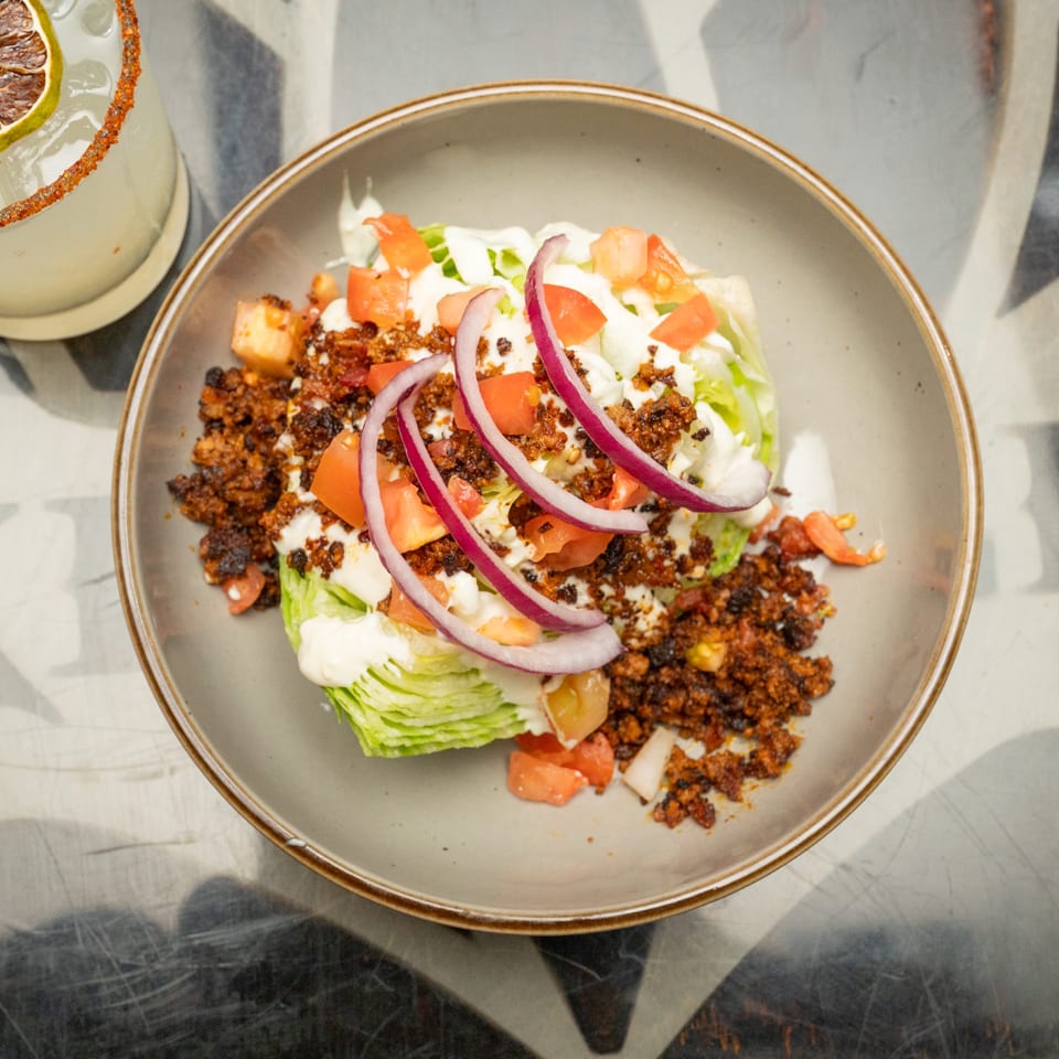 Bowl of chopped lettuce topped with seasoned meat, tomatoes, red onions, and creamy dressing beside a chili-rimmed citrus drink.