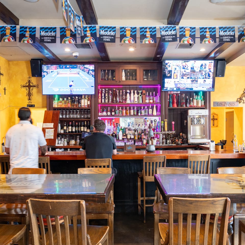 Colorful restaurant bar stocked with liquor bottles, multiple televisions showing sports, Modelo banners, and seated patrons.